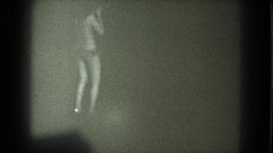 Adult woman appears partially undressed against a dim wall in soft 1975 8mm footage from supplied San Francisco metadata.