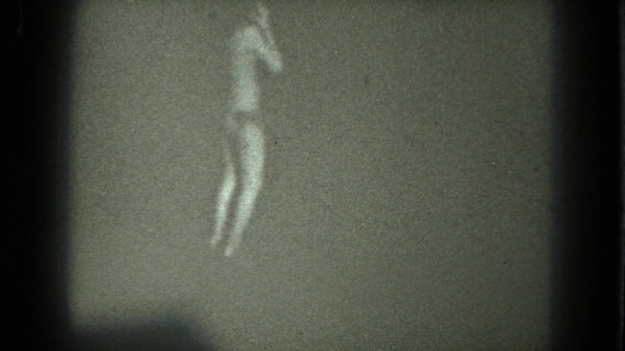 Adult woman in a light bikini and heels dancing against a plain dark backdrop.