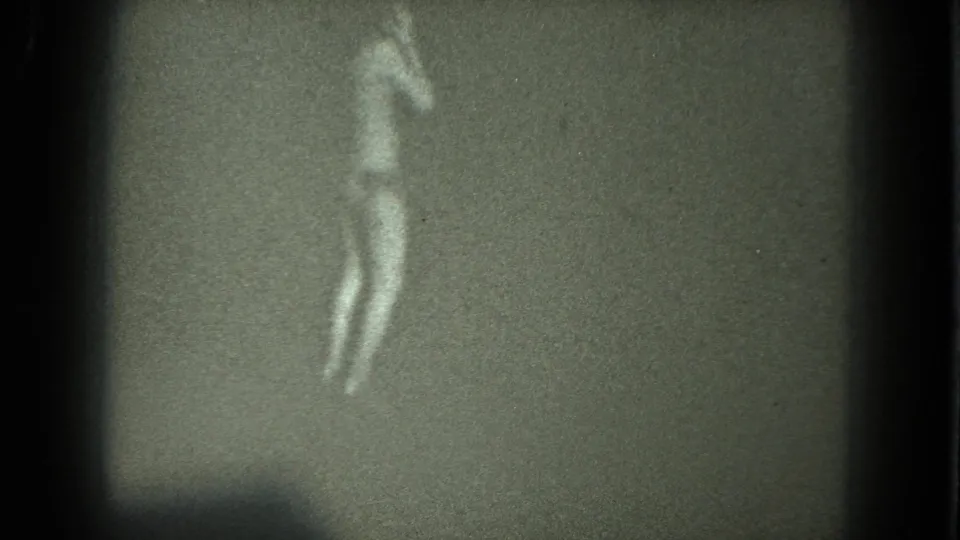 Grainy 1975 8mm frame of an adult woman in a light bikini and heels dancing alone in a dim, plain setting.