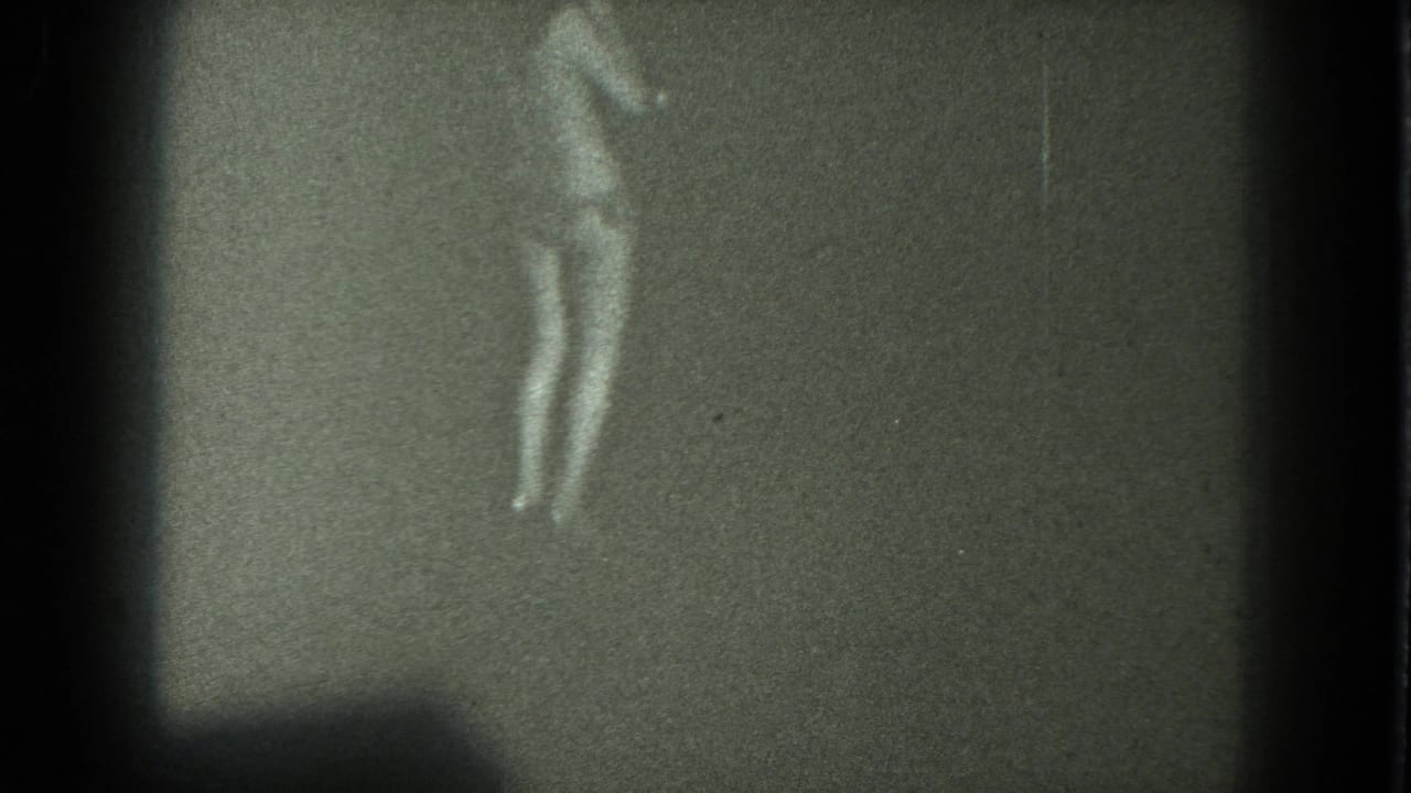 Soft black-and-white 8mm frame showing an adult woman mid-dance in bikini and heels, with no visible audience or setting details.