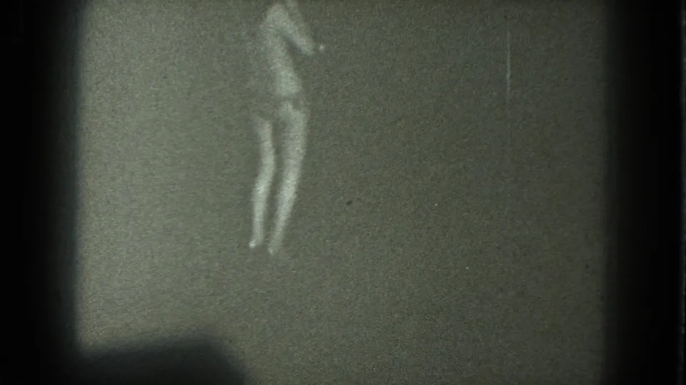 Soft black-and-white 8mm frame showing an adult woman mid-dance in bikini and heels, with no visible audience or setting details.