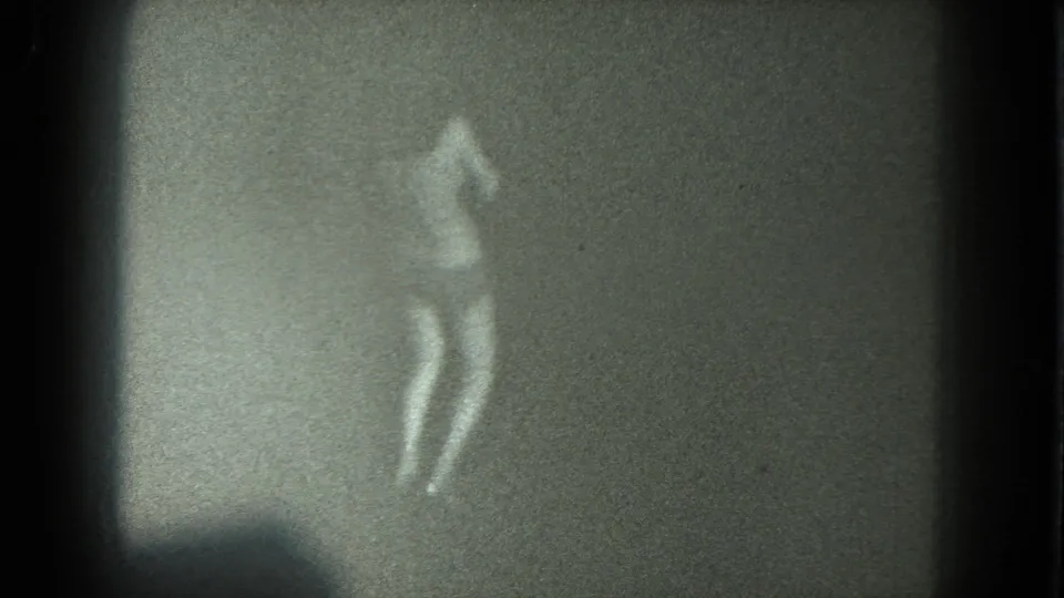 Grainy black-and-white 8mm frame of an adult woman in bikini and heels standing mid-motion against a plain backdrop.