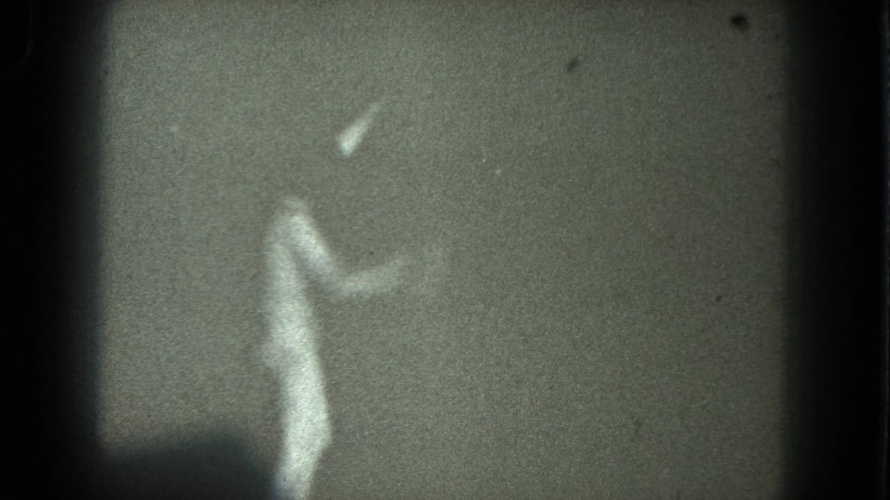 Soft, grainy silhouette of a female dancer against a plain wall with one arm lifted.