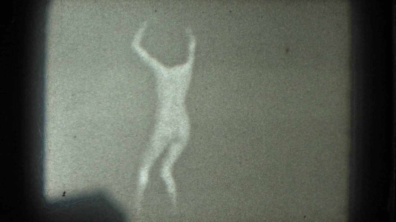 1975 8mm San Francisco frame of a female dancer with arms overhead, rendered as a pale figure against a plain wall.
