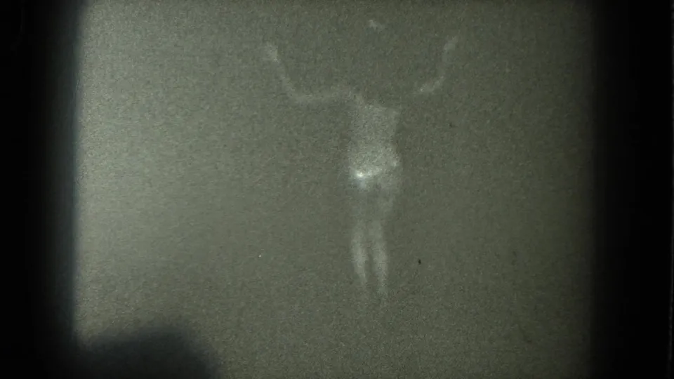 A centered adult figure appears in soft focus, with faint body outline and surrounding 8mm grain in the dim scene.