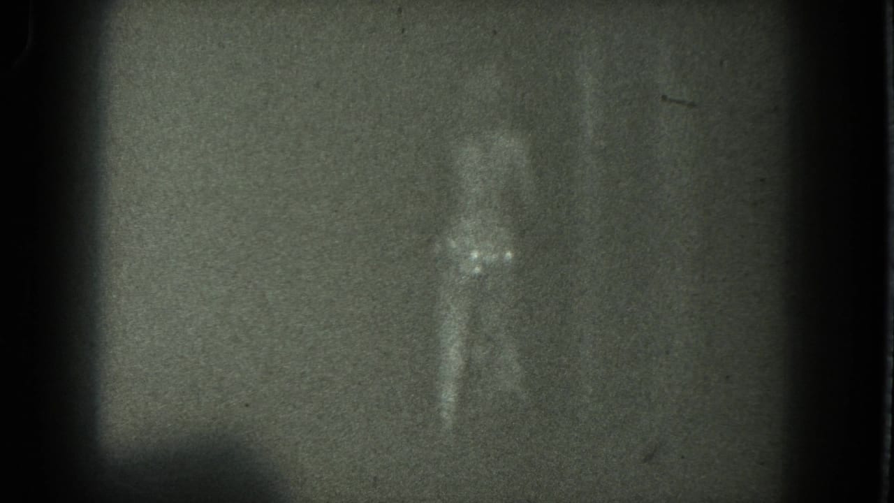 Grainy frame of a woman dancing with one arm raised in low light.
