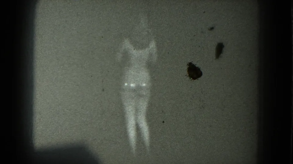 A scantily clad adult woman stands in harsh low light on a dark stage, with 8mm grain and frame dirt visible.