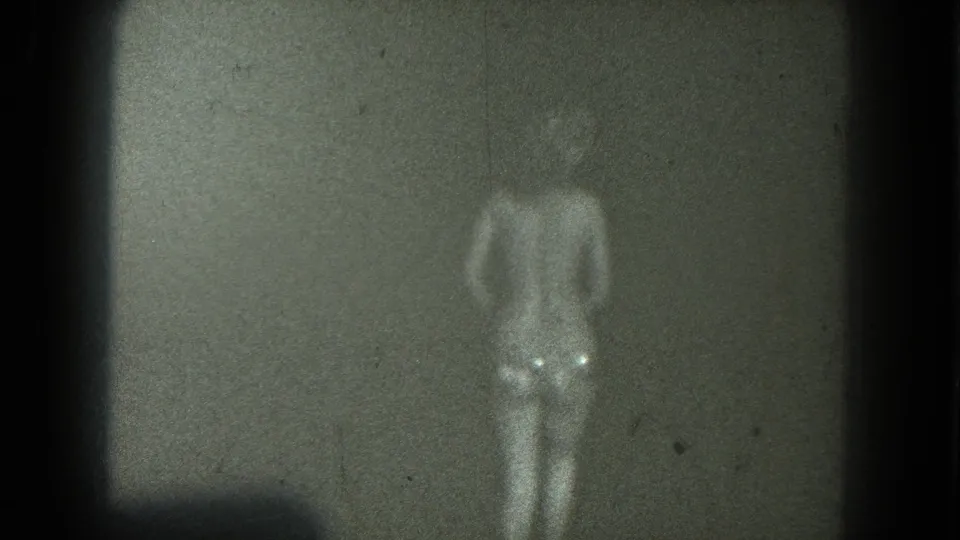 The adult performer appears front-facing and softly overexposed against a plain dark stage background in 8mm footage.