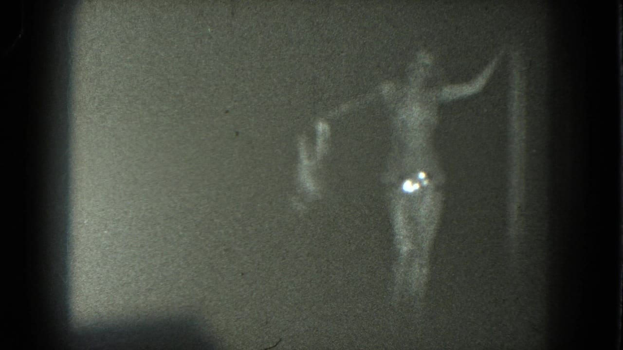 In 1975 San Francisco 8mm footage, an adult performer in a long skirt and bare upper torso poses with one arm raised against a plain backdrop.