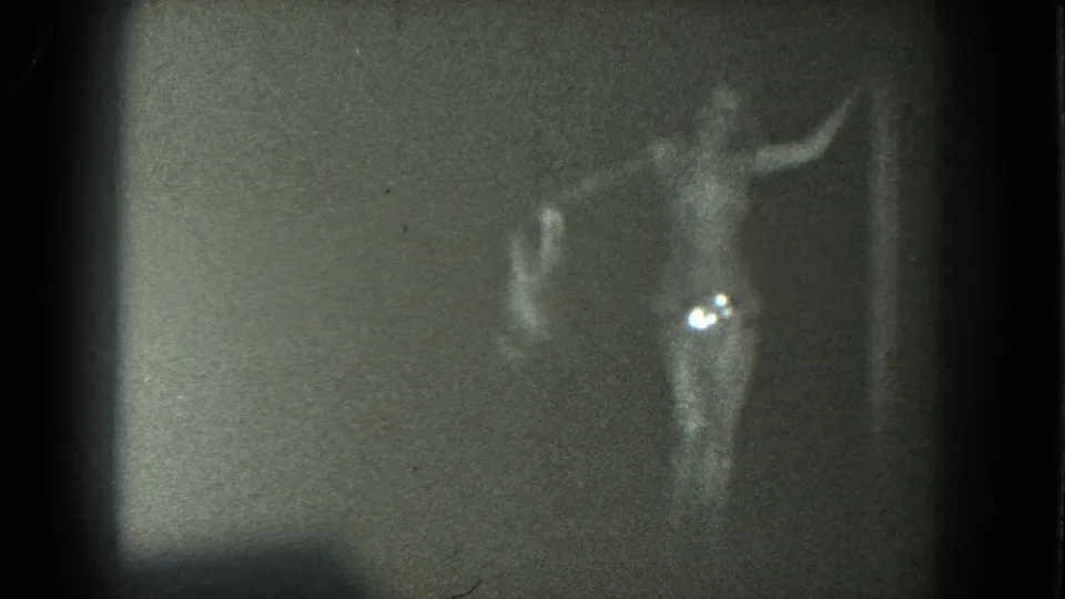 In 1975 San Francisco 8mm footage, an adult performer in a long skirt and bare upper torso poses with one arm raised against a plain backdrop.