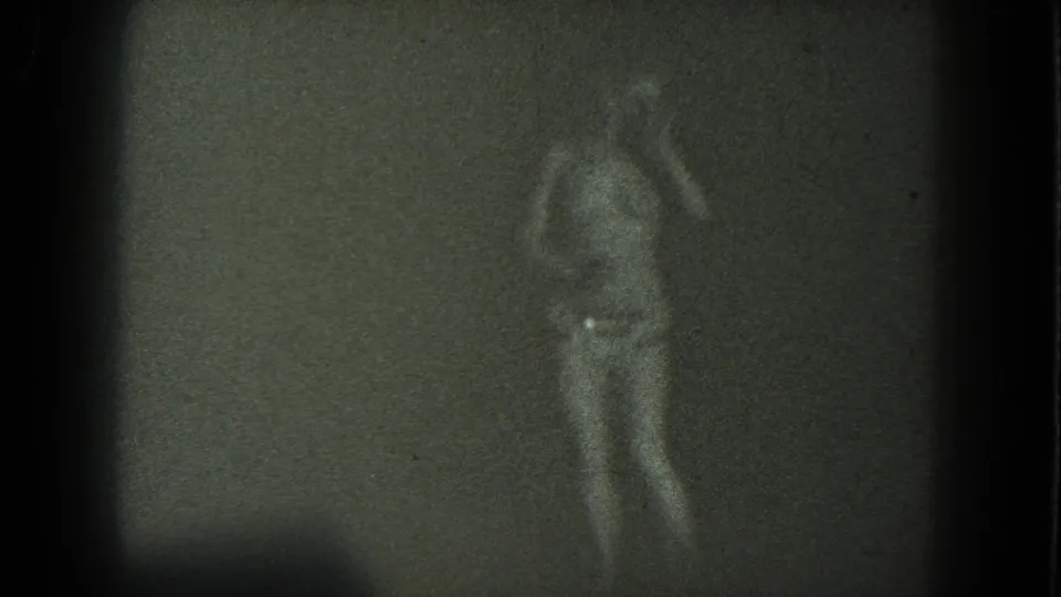 A grainy 1975 San Francisco frame shows an adult dancer in profile, arms lifted, wearing a long skirt against a bare wall or screen.