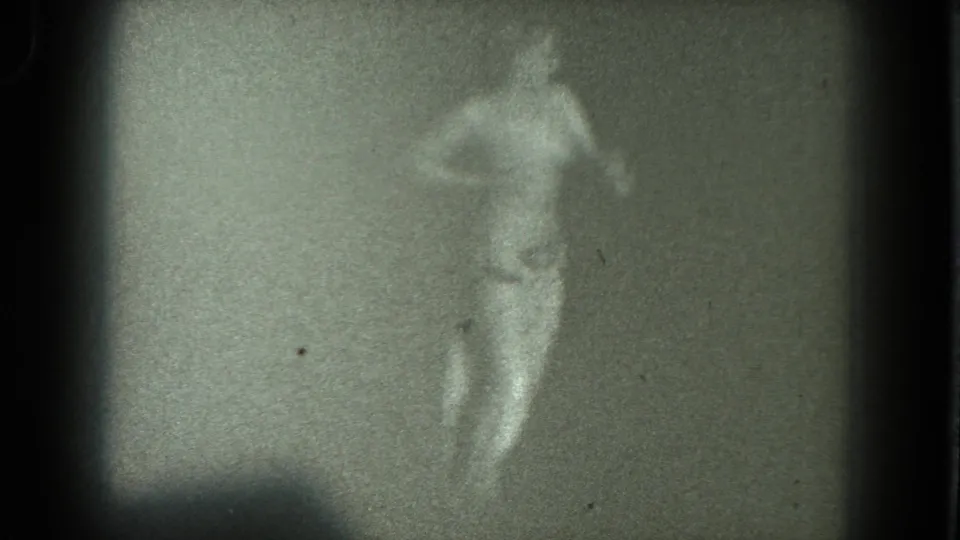 The 8mm frame shows an adult performer mid-gesture, partly blurred by motion, with a long skirt and bare upper torso in dim light.