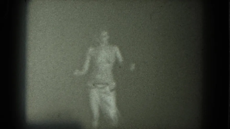 An adult dancer faces the camera near the end of the 1975 San Francisco 8mm clip, arms extended slightly, with a plain backdrop behind them.