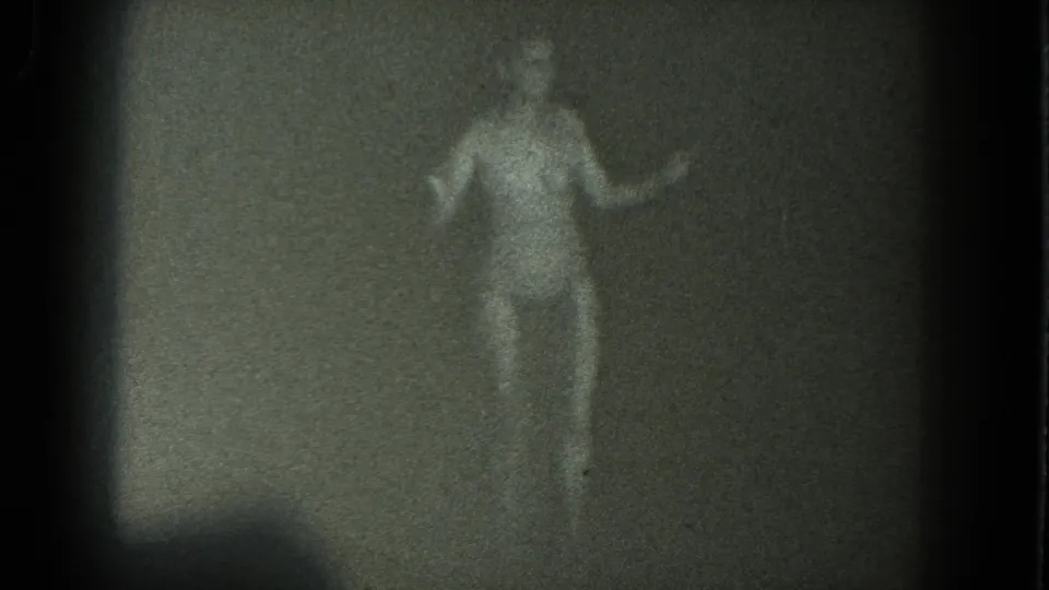 An adult woman in a two-piece bathing suit faces the camera during a dim 1975 San Francisco home-movie dance moment.
