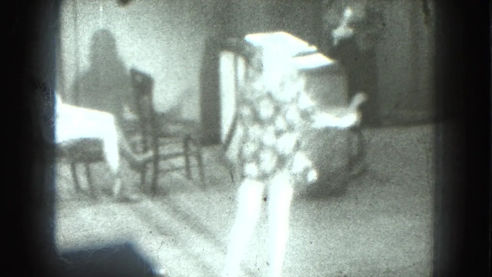 Two adult women dance in a private interior with chairs and a table, seen in grainy 1975 San Francisco 8mm footage.