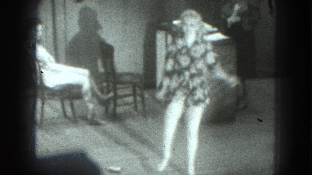 In a grainy 1975 San Francisco 8mm frame, an adult woman in a patterned dress performs in a dim interior with chairs and seated figures.