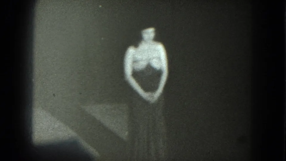 A grainy 1975 San Francisco 8mm frame shows an adult woman in a dark fitted outfit standing still against a plain backdrop.