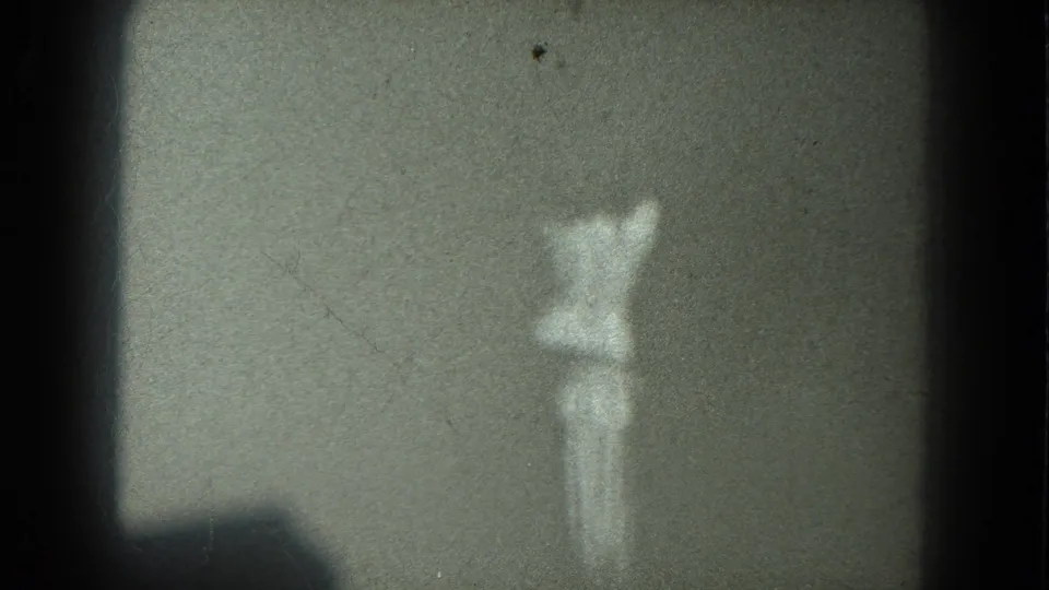 A soft, low-exposure 8mm frame shows the dancer from behind against a plain dark wall, with scratches and frame-edge shadowing.