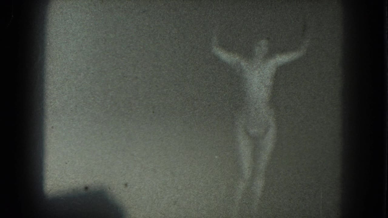 1975 San Francisco 8mm frame showing a faint topless adult woman with arms raised in a dim, grainy scene.