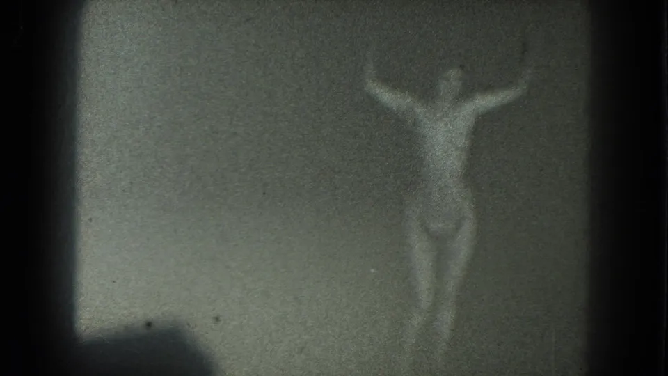 1975 San Francisco 8mm frame showing a faint topless adult woman with arms raised in a dim, grainy scene.