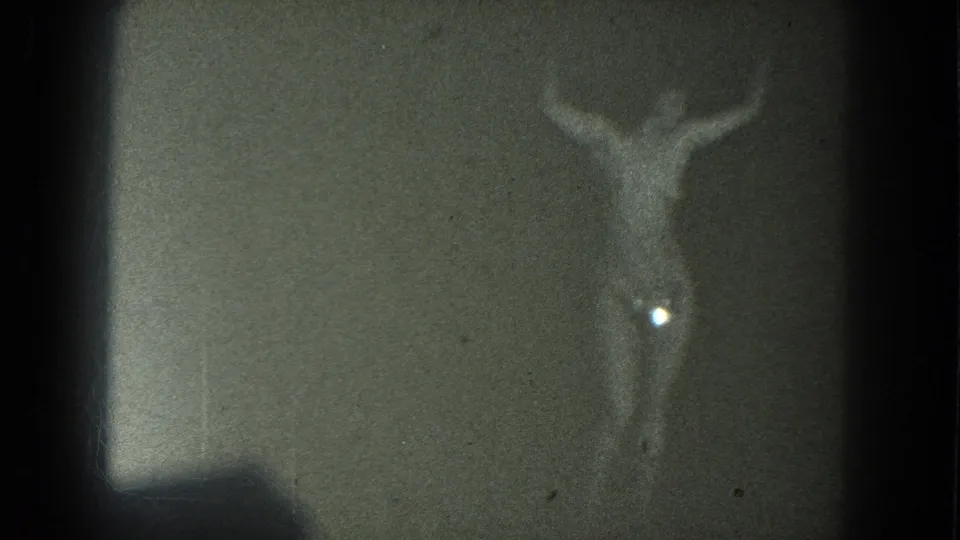 1975 San Francisco 8mm frame with a dim topless adult performer, raised arms, and a bright flare near her side.