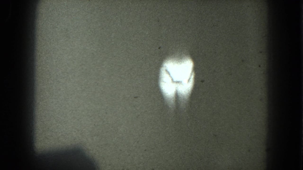1975 8mm frame from metadata-described San Francisco footage showing a bright, indistinct dancer-like silhouette in a dark projected frame.