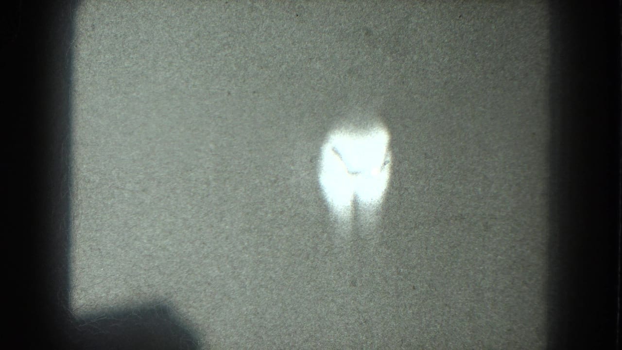 A bright, soft-edged silhouette appears near the upper center of a grainy 8mm frame, with no identifiable face or setting visible.