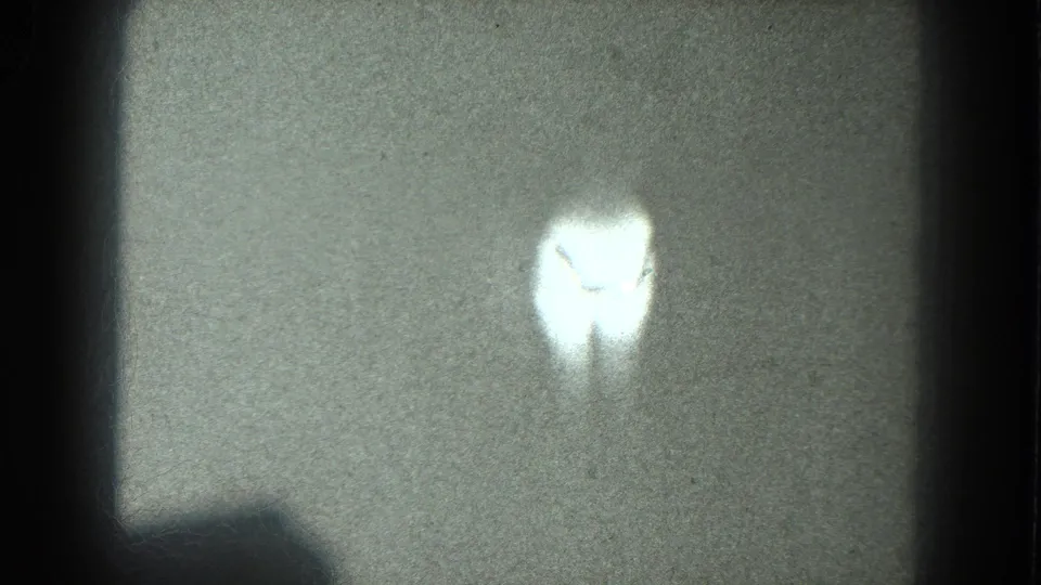 A bright, soft-edged silhouette appears near the upper center of a grainy 8mm frame, with no identifiable face or setting visible.