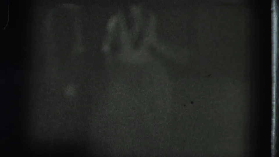 A soft, shadowed silhouette appears within a dark grainy 8mm frame, bordered by a visible vertical film edge at right.