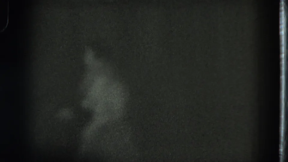 The performer appears in profile during a turn, with a grass skirt and upper-body motion visible in the dark 8mm frame.