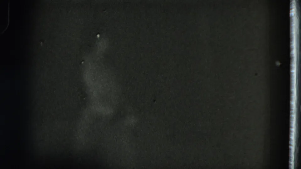 A dim home-movie frame shows the dancer lower in the shot, with the grass skirt shape and body outline faintly visible.