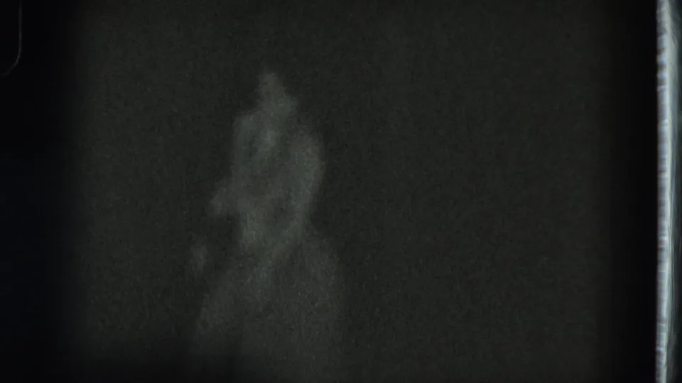 Very dark 1975 San Francisco 8mm frame with a barely visible performer standing against a dim background.
