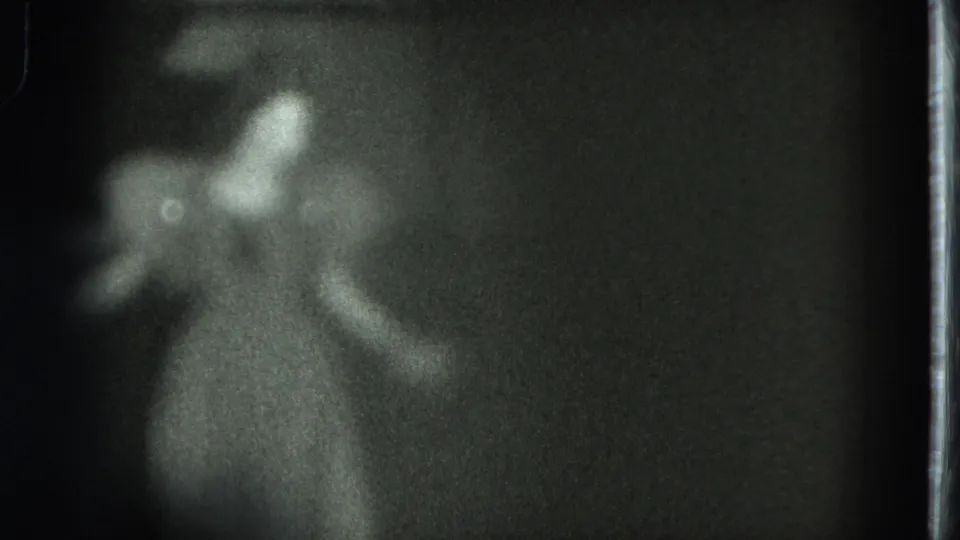 Dark 8mm home-movie frame showing the performer lower in the composition, with a pale dress and indistinct arm movement.