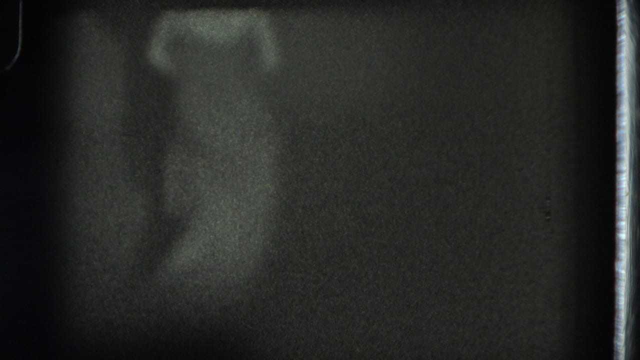 A grainy 8mm frame from the stage performance shows the dancer partly turned, with the body softened by low light and motion.