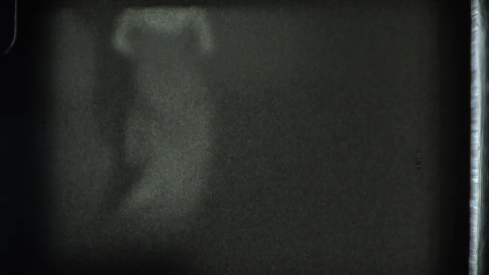 A grainy 8mm frame from the stage performance shows the dancer partly turned, with the body softened by low light and motion.