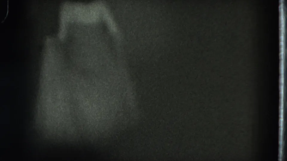The dancer appears lower in the frame, wearing a long dress as the dim stage light catches the figure against a dark background.