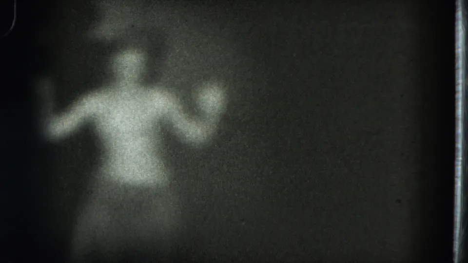Low-light 8mm frame shows an adult woman in light clothing with arms lifted, surrounded by darkness and grain.