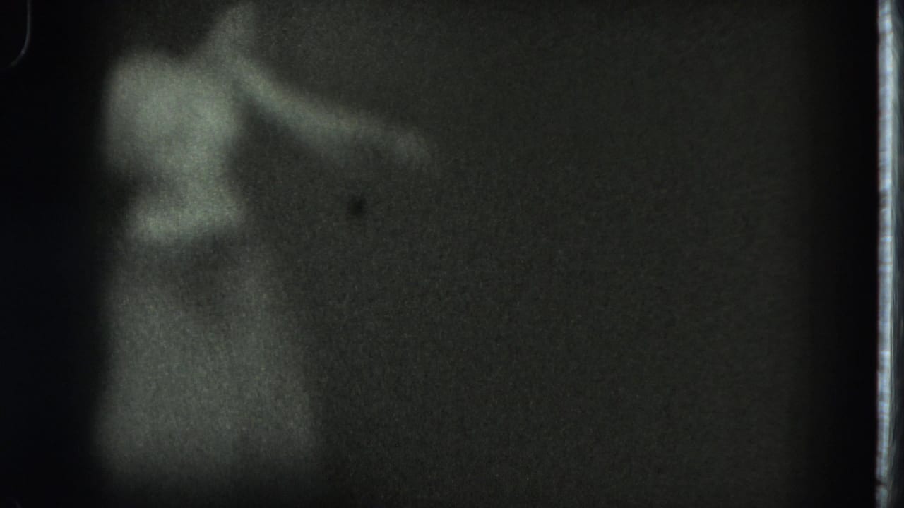 Soft black-and-white 1975 San Francisco home-movie frame showing a blurred adult figure with an arm raised in a dark room.