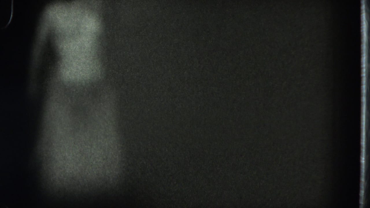 Grainy 1975 San Francisco home-movie frame with a faint adult figure standing or dancing in low light.
