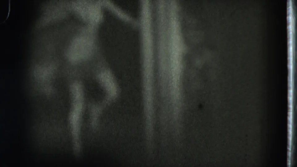 Grainy 1975 San Francisco home-movie frame with a blurred adult dancer near a bright vertical edge in low light.