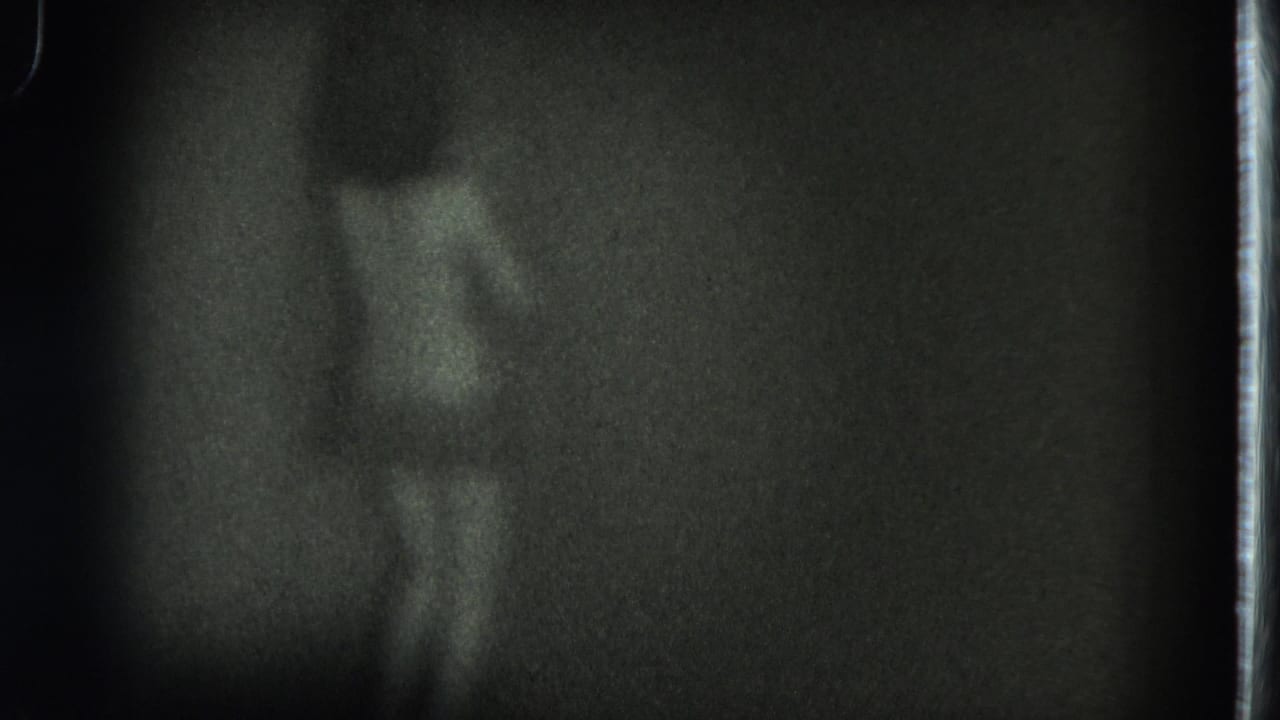 Dim 1975 San Francisco 8mm frame showing a blurred adult figure from behind against a dark interior background.