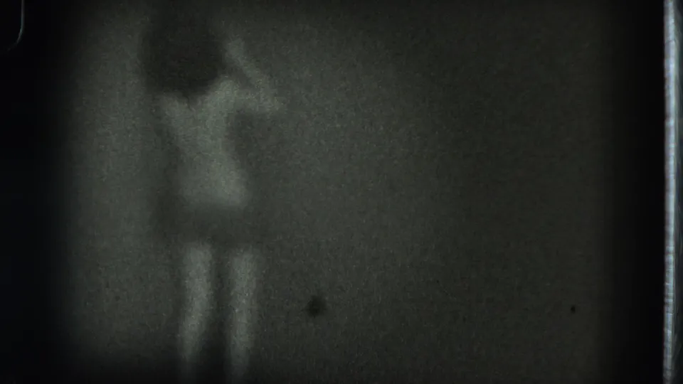 1975 San Francisco 8mm frame showing a blurred topless adult figure from behind in a dark interior.