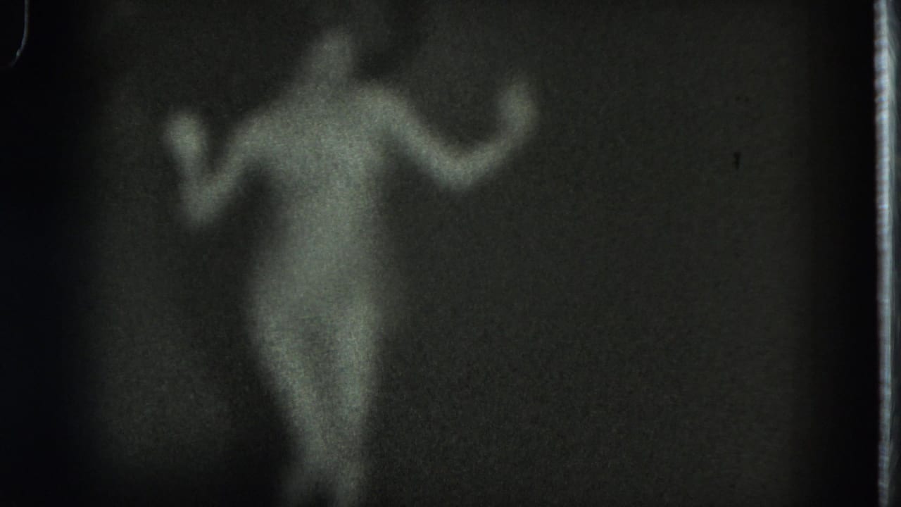 Nude adult woman seen softly in low light with both arms lifted.