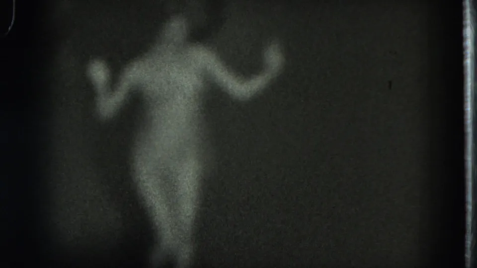 Grainy 1975 8mm frame from San Francisco showing a nude adult woman with arms raised while dancing indoors.