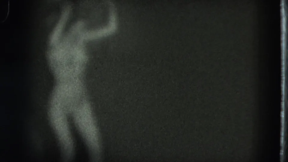 Grainy 1975 8mm frame from San Francisco showing a nude adult woman turning or swaying in a dim interior.