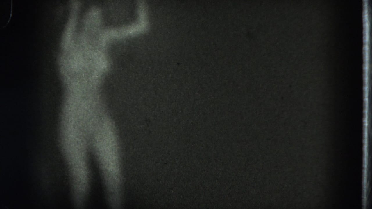 Soft black-and-white 8mm frame of a performer partly emerging from darkness on a stage in the supplied 1975 San Francisco clip.