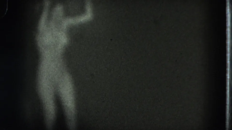 Soft black-and-white 8mm frame of a performer partly emerging from darkness on a stage in the supplied 1975 San Francisco clip.