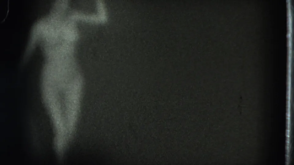 Grainy black-and-white frame showing the performer in profile, partly lit and partly obscured by darkness and film-edge artifacts.