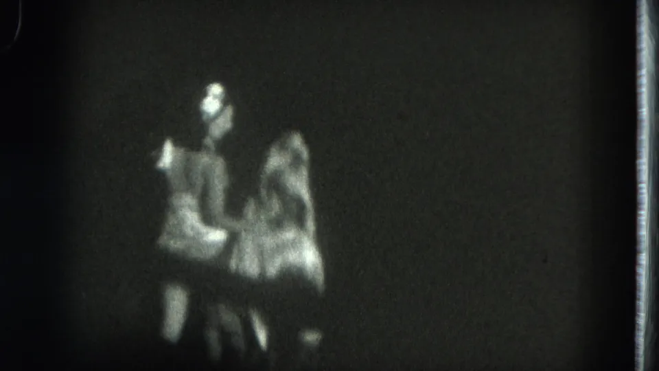 Black-and-white 8mm frame with blurred stage figures, including a performer with raised fabric or costume material under low light.