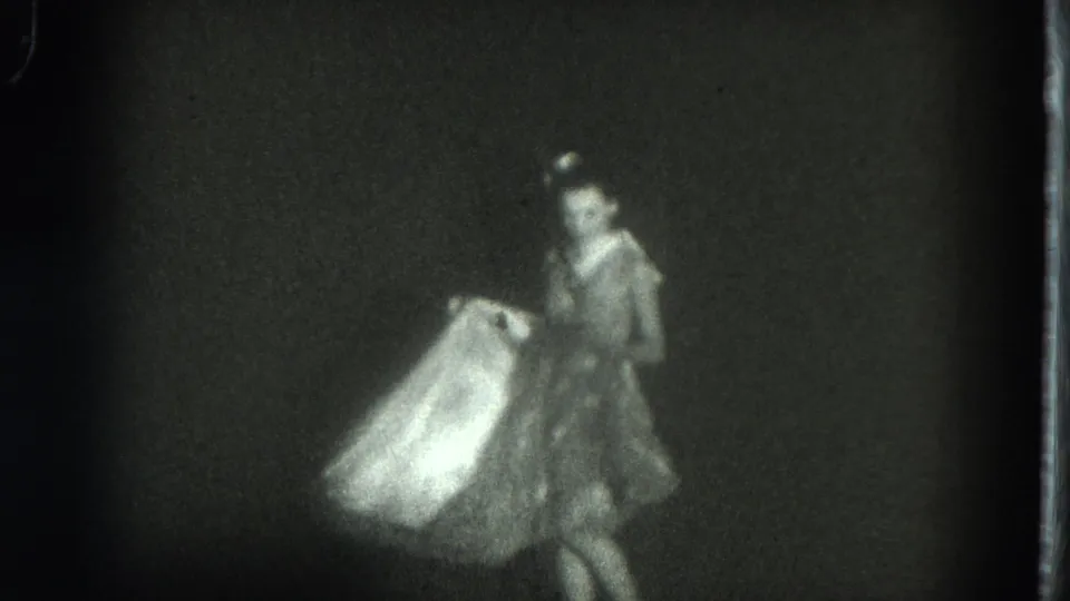 Grainy 8mm frame of a woman on a dark stage holding or moving a pale flowing garment in the 1975 San Francisco performance clip.